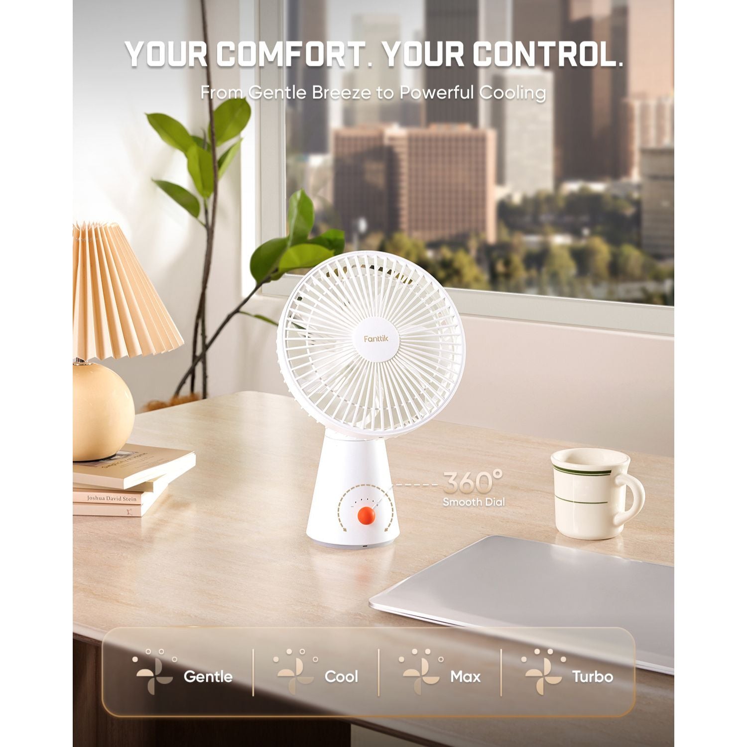 A small white personal fan on a wooden desk in a bright room, with a window view of a city skyline, a lamp, books, a mug, and a laptop. Text at the top reads 'YOUR COMFORT.