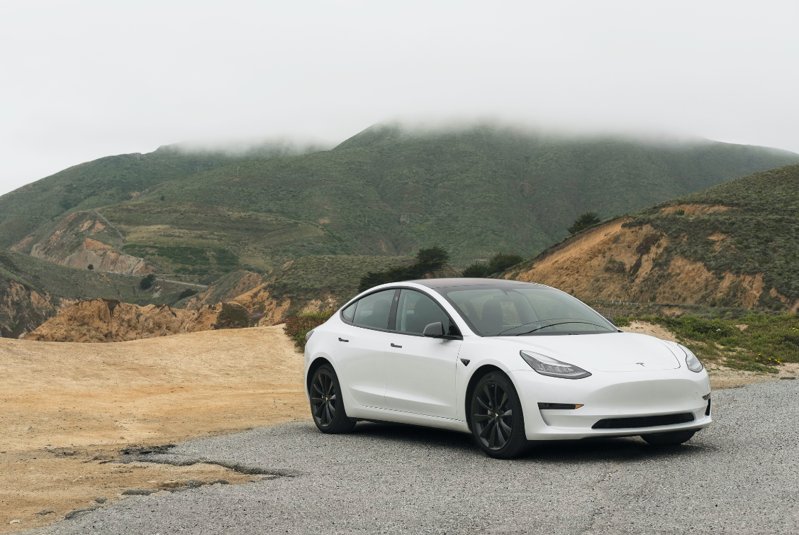 A white Tesla Model 3 sits on a roadside pull-off, framed by fog-shrouded green and brown coastal hills