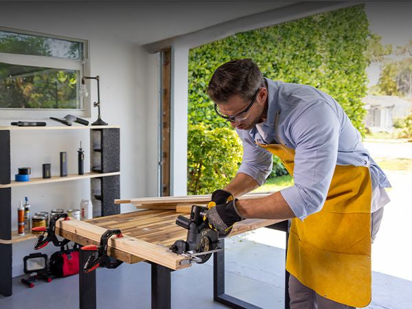 A man uses a Cordless Circular Saw on a woodworking table inside a bright garage workshop with greenery visible outside.