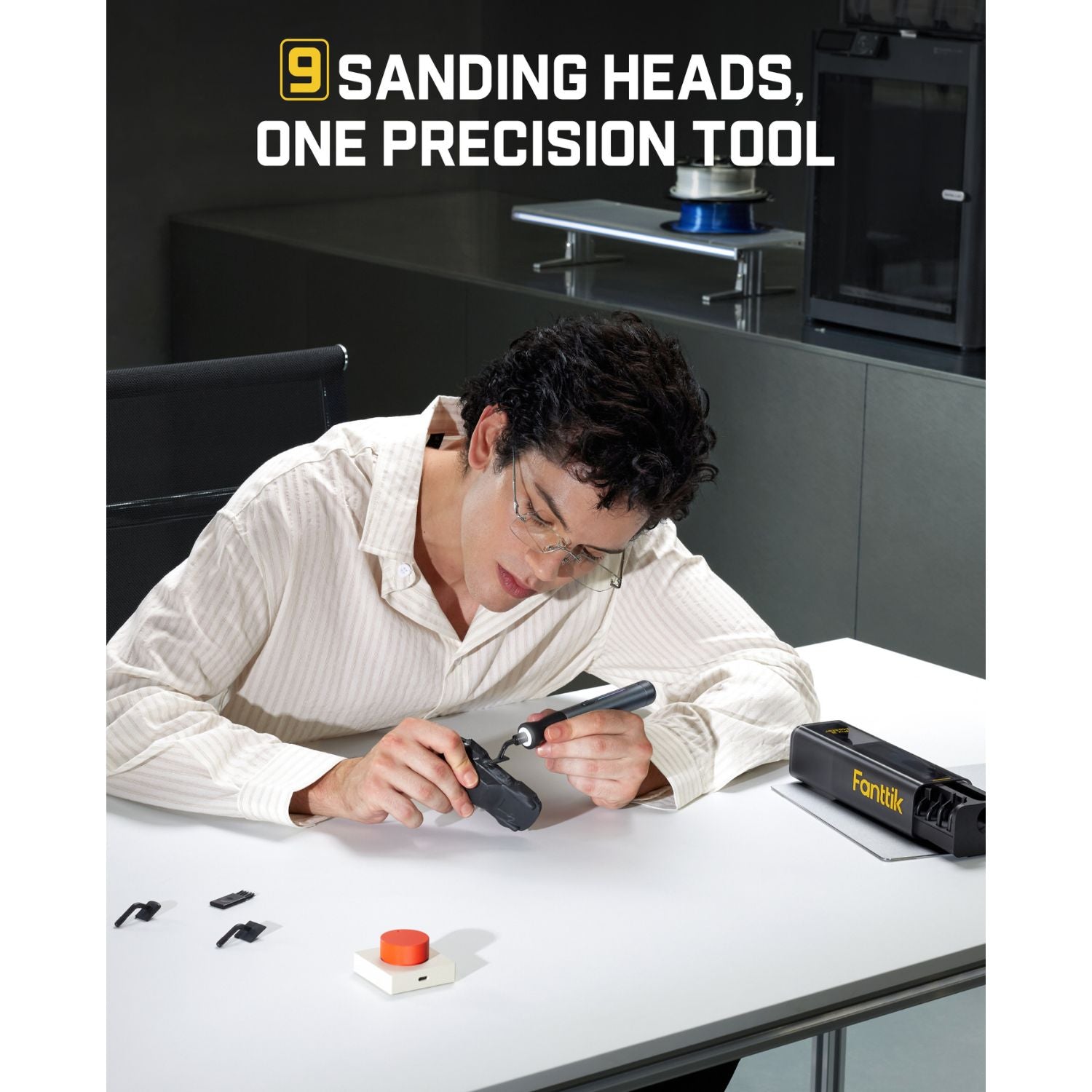 A man in a white shirt leans over a white table, using a handheld Detail Sander. On the table are small black accessories and a red button, with a black branded case nearby.