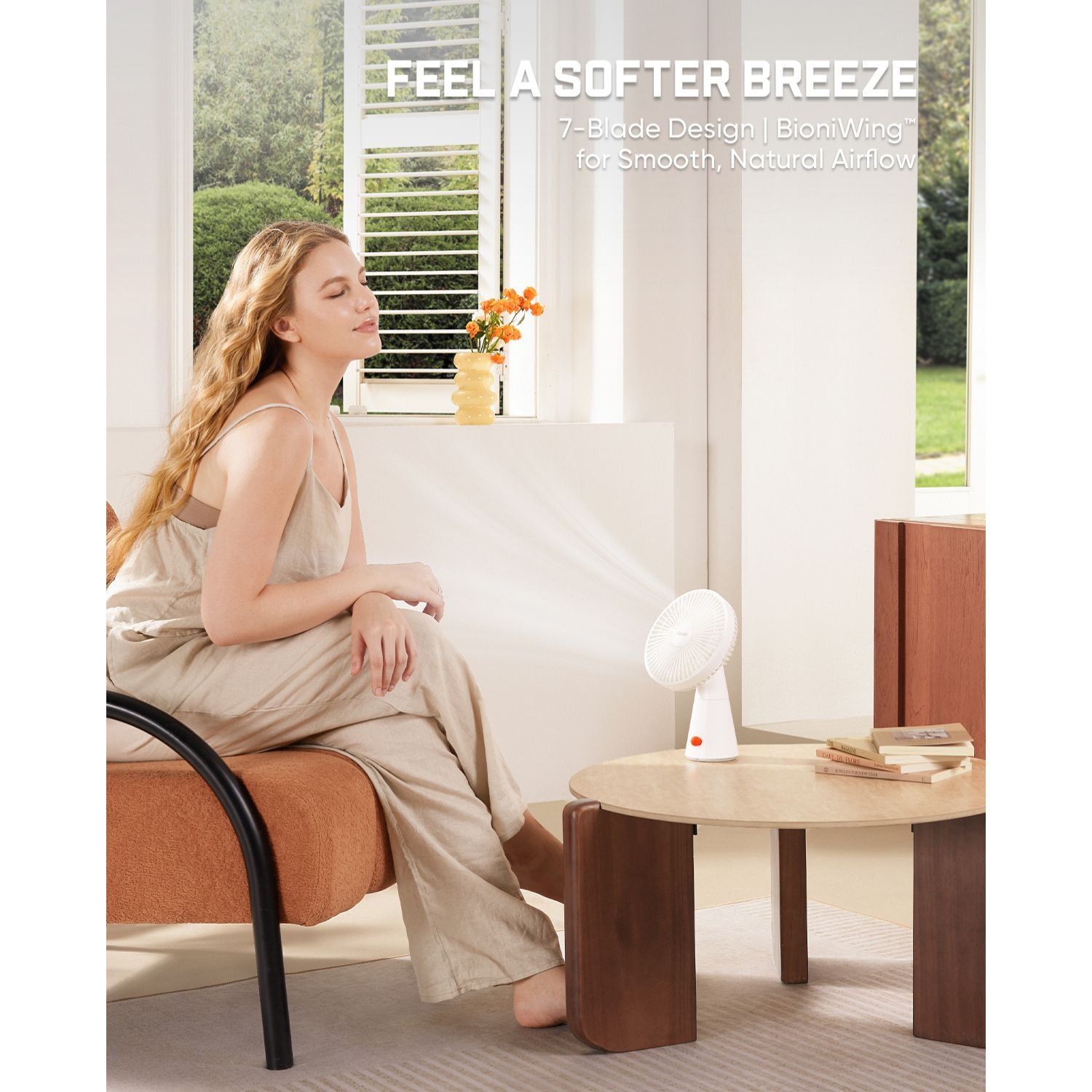 A woman in a beige slip dress sits on an orange upholstered chair beside a small white fan placed on a round wooden table in a bright room. Text at the top reads, 'FEEL A SOFTER BREEZE' with smaller copy about a 7-blade design and biomimicry for smooth, natural airflow.