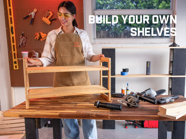 A woman wearing safety glasses and a tan apron stands behind a wooden workbench in a workshop, holding a two-tier wooden shelf. The background shows a pegboard with tools, shelves, and a window.