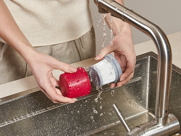 A person washing a red cylindrical filter cartridge with water running from a stainless steel faucet into a sink.
