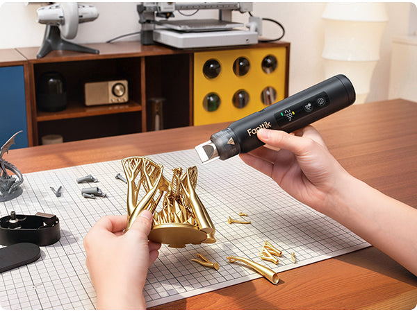 A person uses a black Fanttik H3 Master cordless heat gun  to print small gold-colored components on a white gridded cutting mat at a wooden desk, with a workshop environment in the background.