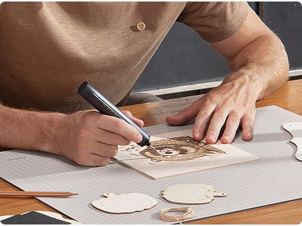 A person using a black T2 Max Cordless Soldering Iron to draw on a small wooden card, with wooden cutout pieces and string on a table mat nearby.