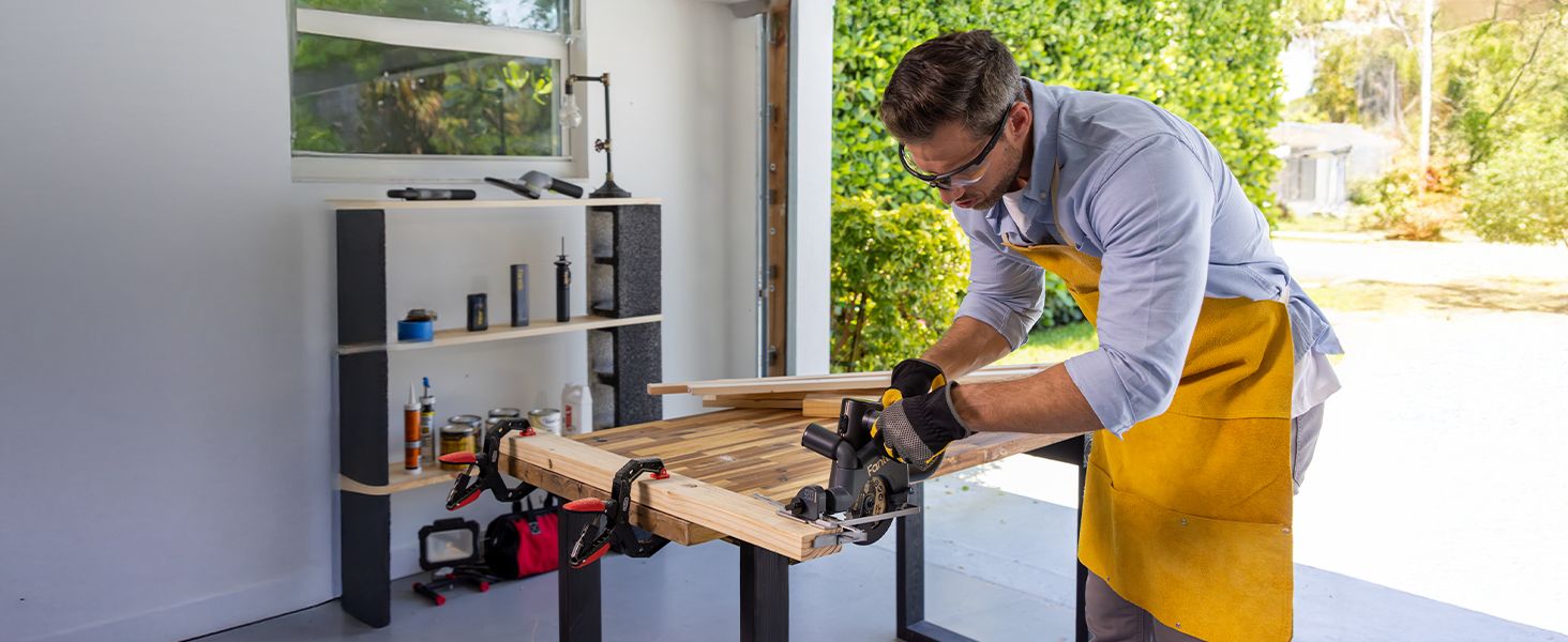 A man works at a woodworking table in a bright garage or workshop, using a Cordless Circular Saw on a wooden board.