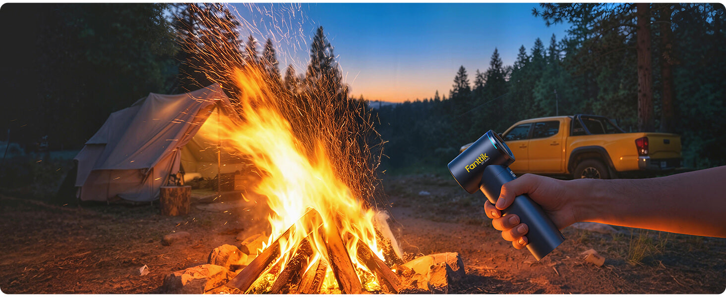 Person holding a Fanttik electric air duster near a campfire at dusk with a tent and truck in the background.