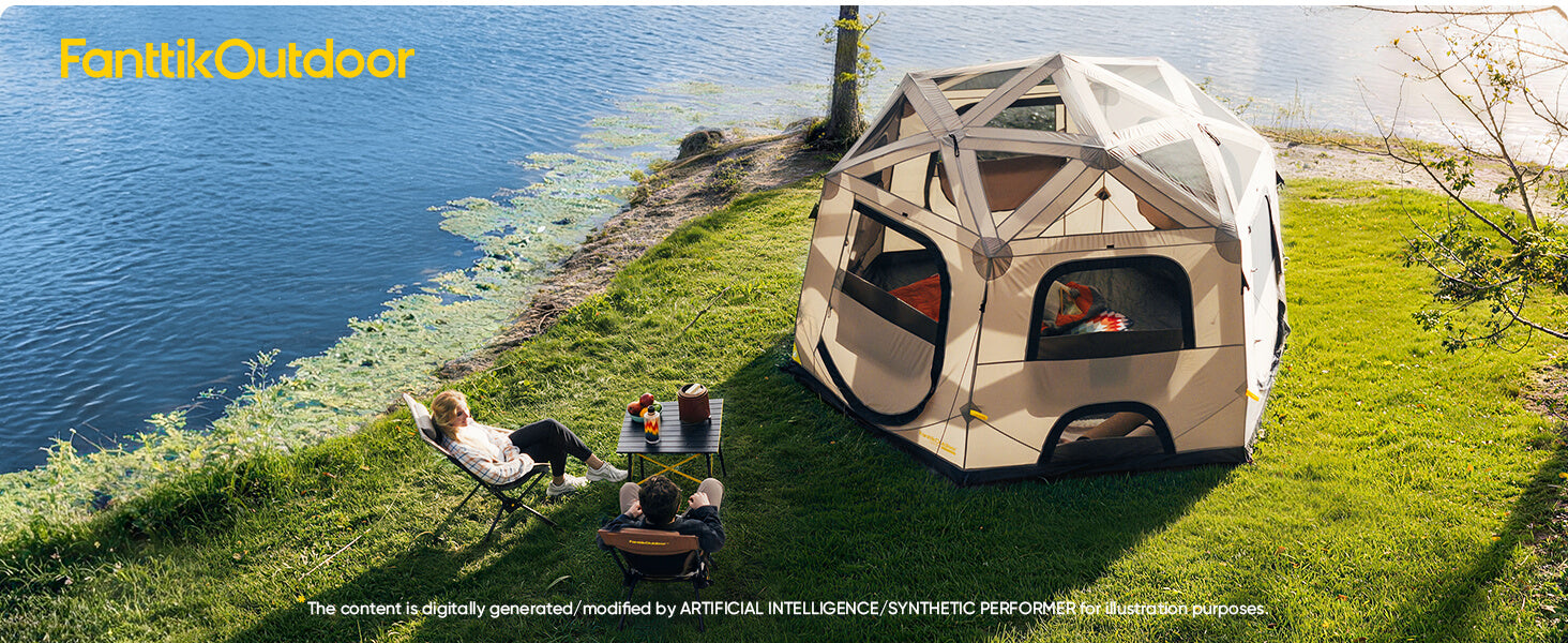 A large geodesic-style outdoor cabin tent in beige and white is pitched on a grassy lakeshore. Two people are relaxing in lawn chairs beside a small table, with the blue lake and green bank forming the background.